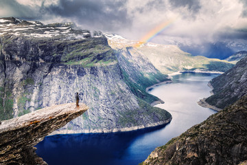 Male climber standing at the edge of cliff Trolltunga looking at rainbow against mountains, dramatic sky and amazing blue lake. Location: beautiful landscape of wild nature in Norway, Scandinavia.