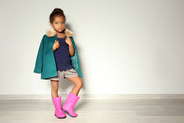 Cute little African American girl in coat against light wall. Fashion concept