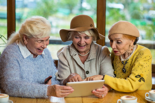 Senior women with tablet. Smiling ladies at cafe table. Looking through the childhood photos.
