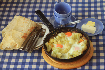 cauliflower and fried eggs in a pan.  Bread, forks, knives, butter, cups around the pan.  A table with a checkered tablecloth