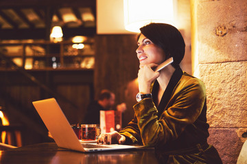 Attractive business woman is smiling aside while sitting with the lectronic devices in a coworking space. Fashion blogger is holding the smartphone while sitting in a cafe with a laptop.