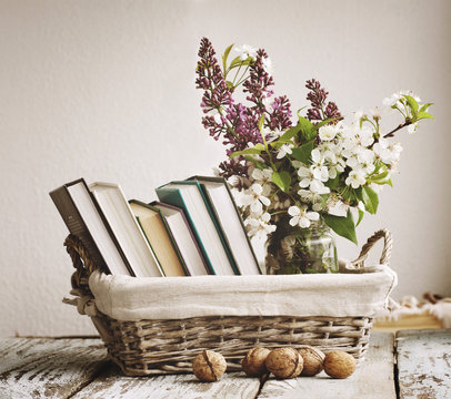 Pile of books and spring flowers of a lilac and apricot in a basket on a wooden surface. Vintage still life.