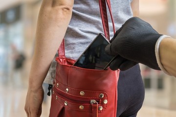 Pickpocket thief is stealing smartphone from red handbag.