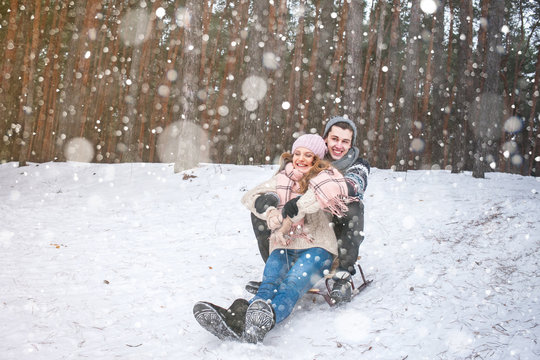 Young couple in winter forest sledding, laughing during snowfall