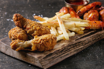 Fast food fried crispy chicken legs and french fries potatoes with salt served on wooden serving board over dark texture background. Space for text. Close up