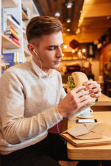 Young handsome man sitting in cafe while holding sandwich.