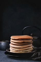 Stack of homemade american ombre chocolate pancakes served on black plate with jug of cream and teapot over black stone texture background.