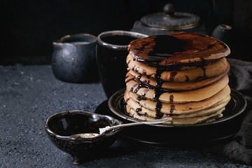 Stack of homemade american ombre chocolate pancakes with carob honey sauce served on black plate with jug of cream and teapot over black stone texture background.