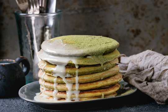 Stack of homemade american ombre green tea matcha pancakes with condensed milk sauce and matcha powder served on gray plate with jug of cream and cutlery over black stone texture background.