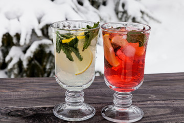 Two glasses of Fresh sweet cold carbonated homemade lemonade with lemon slices, strawberry juice and mint leaves on old dark grey rustic wooden table. Christmas fir branches with snow on background.
