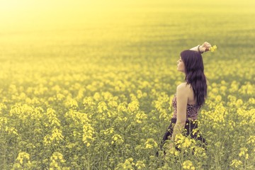 Fototapeta premium Schöne junge Frau im Sommer auf gelber Wiese seitlich von hinten. Das hübsche Mädchen steht in einem weiten Feld voller Raps und genießt den warmen Sonnenschein im Frühling. Rückansicht seitlich