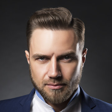 Close-up face of handsome young man with beard and hair style wearing suit and shirt over dark background