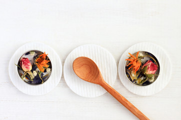 
Containers with dried aromatic medicinal flowers on cotton pads, wooden spoon, empty space white wooden table background, top view set.