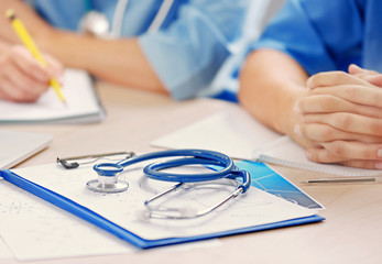 Stethoscope, clipboard on desk and interns on background, closeup