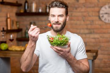 Handsome bearded man in white t-shirt eating salad with tomatoes in the kitchen. Healthy and vegan food concept