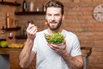 Selbstklebende Fototapeten Handsome bearded man in white t-shirt eating salad with tomatoes in the kitchen. Healthy and vegan food concept © rh2010