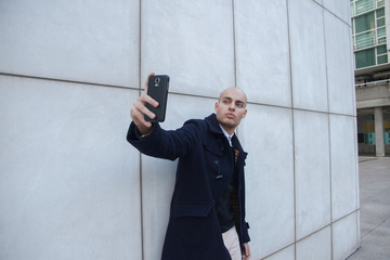 Young handsome bald businessman holding a smart phone doing a selfie leaning against a white wall - technology, business, work concept  