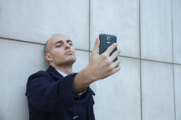 Young handsome bald businessman holding a smart phone doing a selfie leaning against a white wall - technology, business, work concept  