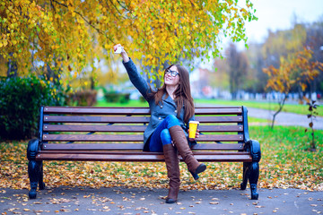 The girl in the jacket makes selfie photo, sitting on a bench with glasses, holding  coffee or tea,  young outdoors, spring  fall, life style, the concept of the city, lifestyle, smile, happy. For soc