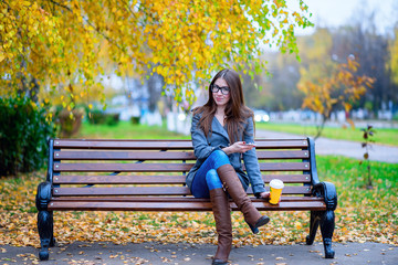 Girl in jacket sitting on the bench with glasses, holding a coffee or tea,  young outdoors, spring  fall, life style, the concept of the city, lifestyle, smile, happy.  the phone in hand.