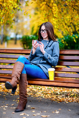 Girl in jacket sitting on the bench with glasses, holding a coffee or tea,  young outdoors, spring ...
