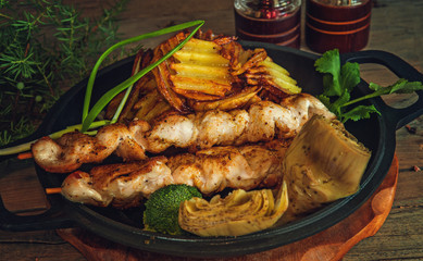 chicken kebabs on wooden skewers with broccoli, artichoke and potato crisps with herbs in a frying pan and a wooden Board