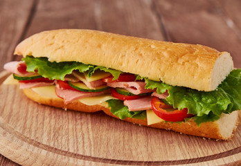 Submarine sandwich with ham, cheese, tomatoes, lettuce, cucumbers and onions on a wooden background