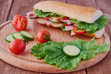 Submarine sandwich with ham, cheese, tomatoes, lettuce, cucumbers and onions on a wooden background