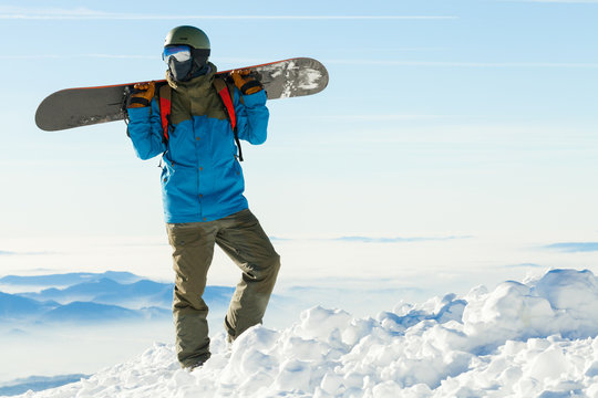 Young snowboarder in helmet standing at the top of a mountain with beautiful sky on background