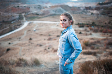 portrait of the attractive, slender, beautiful young Caucasian blonde girl in a jeans jacket. Smiling girl enjoys fine warm sunner weather highly in mountains against the sea
