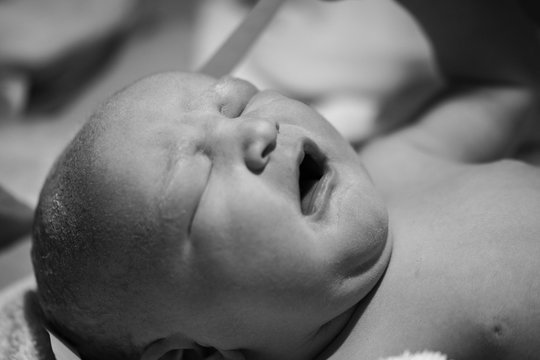 Newborn caucasian baby eyes closed crying naked freshly born black and white being closely examined by paediatric doctors close up of baby head and face in profile the background and body soft focus