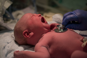 Newborn baby being examined by paediatric doctors moments after birth with stethoscope and gloves checking vital signs in neonatal care crying caucasian female daughter 