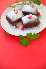 Vertical view of mini chocolate cakes dusted with icing or powdered sugar, topped with fresh raspberries on a plate with tiny ornaments and ivy leaves