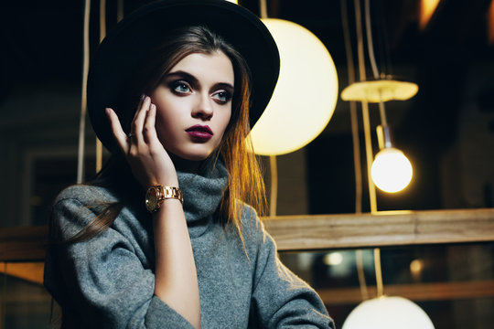 Portrait of a young beautiful fashionable woman touching her face, looking forward. Girl sitting in cafeteria. Model wearing stylish turtleneck and wide-brimmed hat. Close up. Female fashion concept