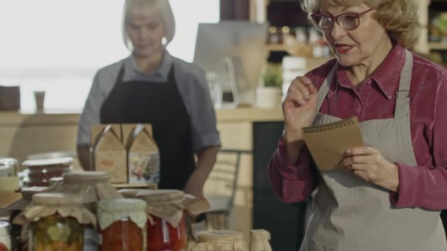 Tilt up of elderly female owner in glasses standing in farmer market counting canned vegetables as her senior colleague stocking products 