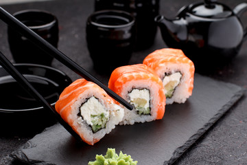 Salmon sushi roll on a stone plate with chopsticks over concrete background.
