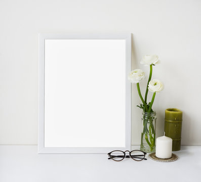 Empty white frame mockup for design presentation, promotional or art content and ranunculus bouquet in glass vase, candles and glasses. Light romantic style concept, minimalism design.