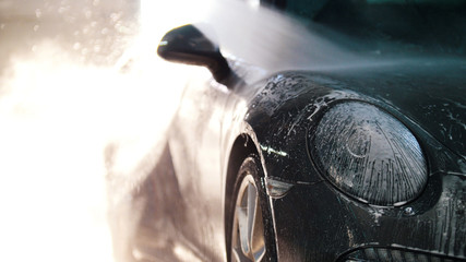 Worker in auto service is washing a luxury car by water hoses, backlight