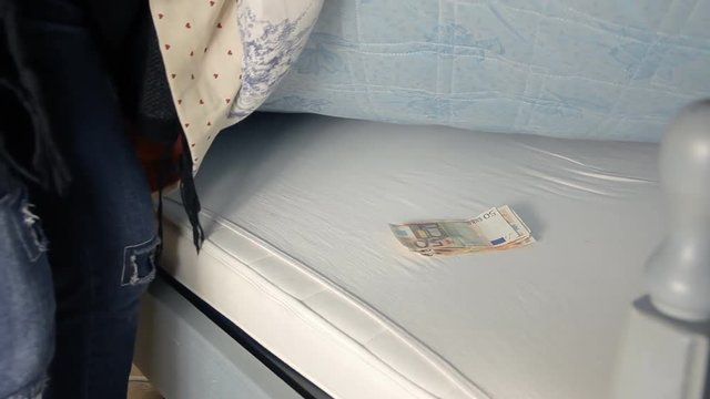 A woman picks a handful of euro banknotes (money) from under the mattress of her bedroom in semi-darkness.
