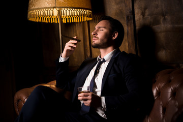 Handsome man drinking whiskey and smoking cigar. Billionaire man resting and relaxing while sitting...