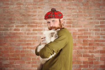 Obraz na płótnie Canvas Young bearded man in plaid cap with fluffy cat on brick wall background