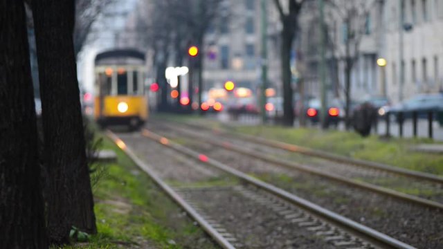 Tram che arriva sfuocato su binario doppio costeggiando strada trafficata sul fondo luci e semafori della citt&agrave;