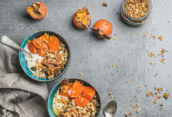 Healthy vegetarian breakfast. Oatmeal, quinoa granola with yogurt, dried fruit, seeds, honey, fresh persimmon in bowls over grey background, top view, copy space. Gluten free, allergy-friendly concept