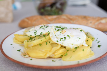 white circular dish with typical Spanish food, sliced fried potatoes and eggs and chopped parsley, with two silver forks and bread on beige tablecloth
