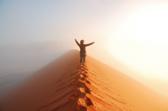 Person standing on top of dune in desert and looking at rising sun in mist with hands up, travel in Africa, Namibia

