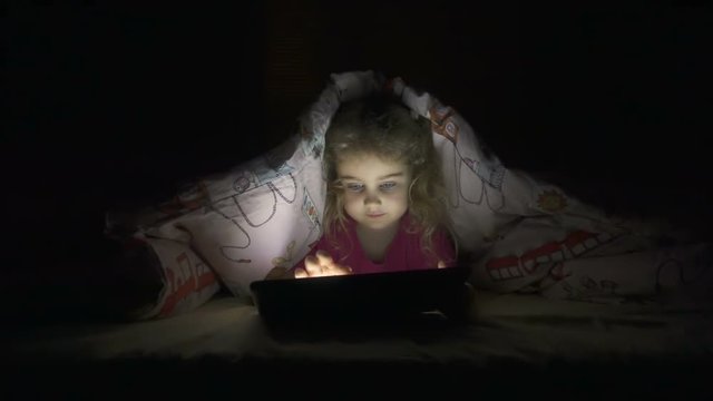 Child Playing on a Tablet at Night. Little Girl Hiding Under Duvet And Use Digital Tablet Device Late After Bedtime.