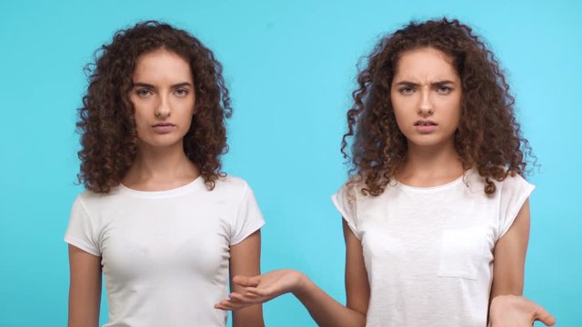 Two embarrassed young Caucasian female curly twins in white t-shirts standing at a loss on blue background