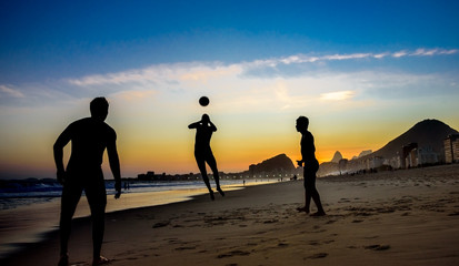 Silhouettes of three men playing beach football on the background of beautiful sunset, Atlantic ocean, Dois Irmaos Mountain and Pedra da Gavea at Copacabana beach, Rio de Janeiro, Brazil