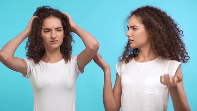 Two embarrassed young Caucasian female curly twins in white t-shirts standing at a loss on blue background in slowmotion