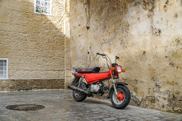 Classic old red scooter parked next to wall in tight street.
